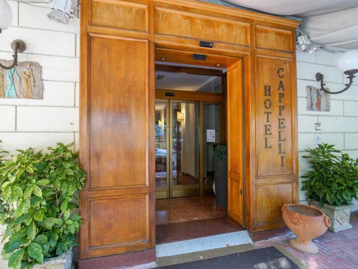 Indgang til Hotel Cappelli med store trædøre, potteplanter på begge sider og en glasdør, der fører ind til lobbyen.