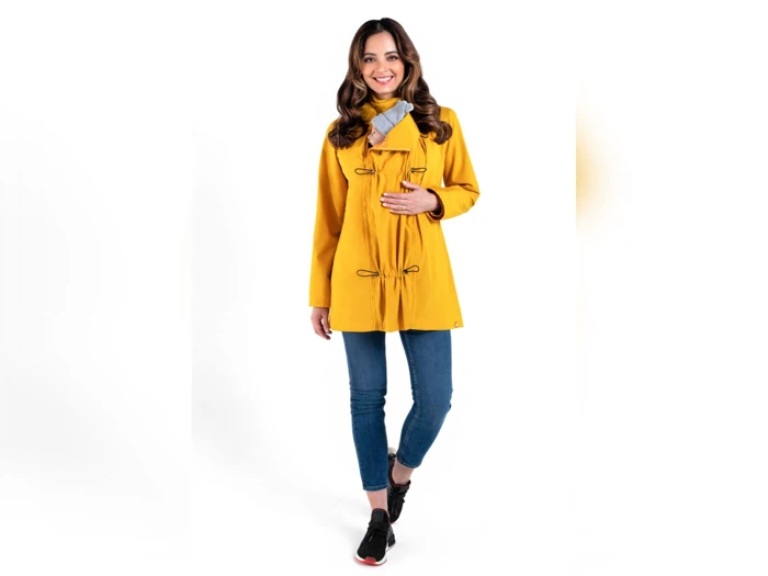 En kvinde iført gul frakke, blå jeans og sorte sneakers står smilende med den ene hånd på sin frakke mod en hvid baggrund.