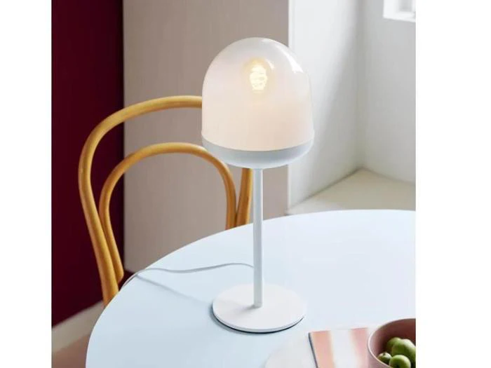 En moderne hvid bordlampe med en kuppelformet skærm af matteret glas står på et rundt hvidt bord ved siden af en træstol.