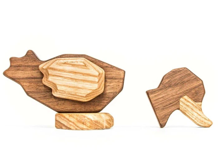 To minimalistiske dyrefigurer i træ, den ene ligner en kylling med lagdelte dele, og den anden ligner en kylling, begge fremstillet af forskellige nuancer af træ.