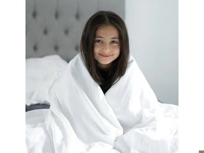 En ung pige med langt brunt hår sidder på en seng, svøbt i et hvidt tæppe, og smiler blidt til kameraet i et lyst soveværelse.