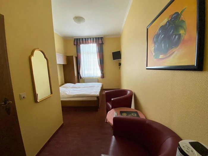 Et lille hotelværelse med en dobbeltseng, et vindue med stribede gardiner, to røde stole, et vægspejl, et tv og et maleri af vindruer på en gul væg.