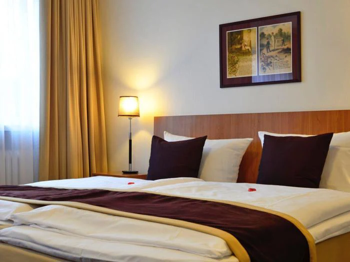 En pænt redt hotelseng med hvidt sengetøj, brune og gyldne detaljer, to puder, en sengelampe og indrammet kunst over hovedgærdet ved siden af et vindue med beige gardiner.