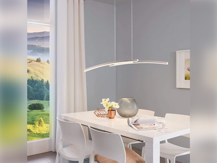 En moderne spisestue med et hvidt bord, fire hvide stole, minimalistisk indretning og en buet pendellampe. Et stort vindue viser en naturskøn udsigt over grønne bakker og blå himmel.