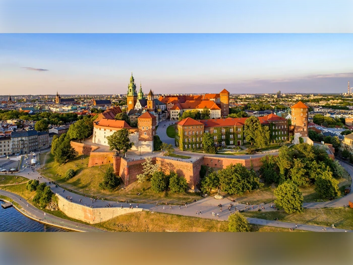 Luftfoto af Wawel-slottet i Kraków, Polen, med bygninger med røde tage, grønne spir og omgivende træer under en klar himmel ved solnedgang.