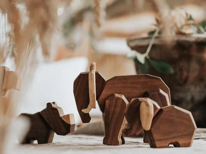 Tre dyrefigurer af træ, bl.a. en elefant og en flodhest, er anbragt på en blød overflade med slørede planter i baggrunden.