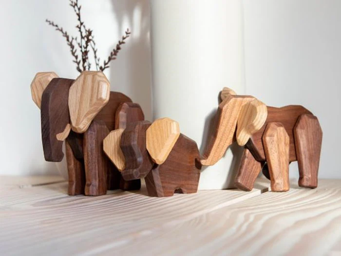 Tre elefantfigurer af træ i forskellige størrelser og træfarver er arrangeret på en lys træoverflade i nærheden af en hvid vase med tørrede grene.