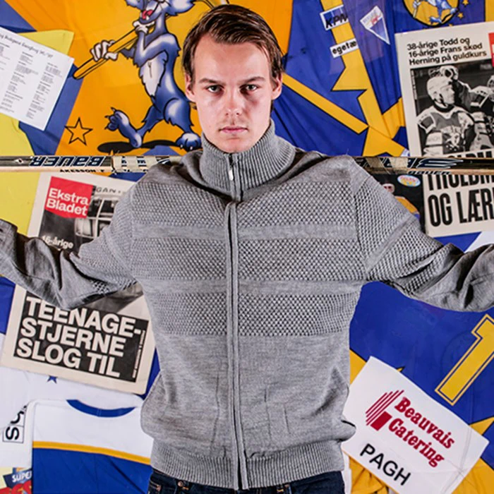 En mand i en grå sweater holder en hockeystav over skuldrene og står foran en farverig baggrund med plakater, avisudklip og tegneseriegrafik.