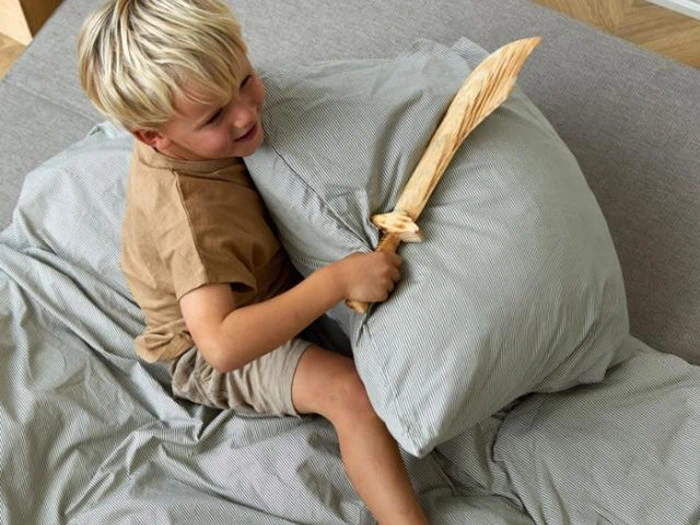 En ung dreng med lyst hår sidder på en seng med et legetøjssværd af træ i hånden og trykker legende på en pude med det.