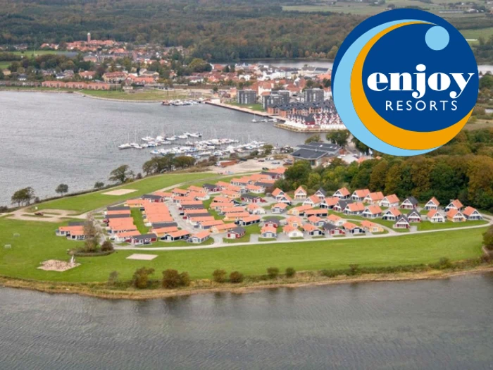 Luftfoto af et kystresort med huse med røde tage nær vandet, træer og havne, med Enjoy Resorts logo i øverste højre hjørne.