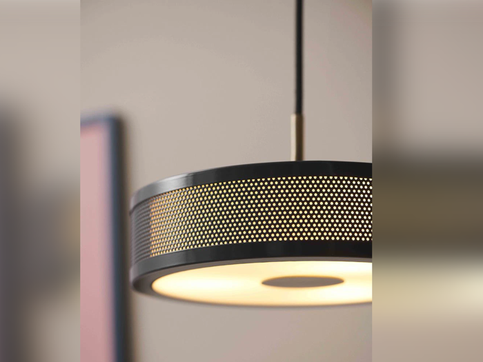 Et nærbillede af en moderne sort pendel med en perforeret metalskærm, der lyser blødt og hænger ned fra loftet mod en neutral baggrund.