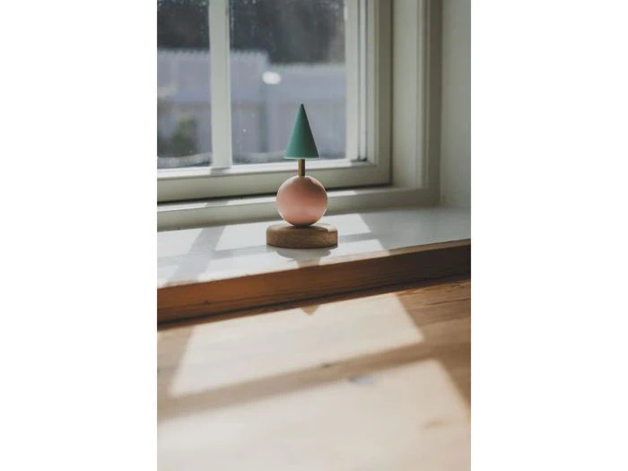 Et lille trælegetøj, der ligner et træ med en grøn kogle på toppen af en lyserød kugle, står i en vindueskarm i naturligt sollys.