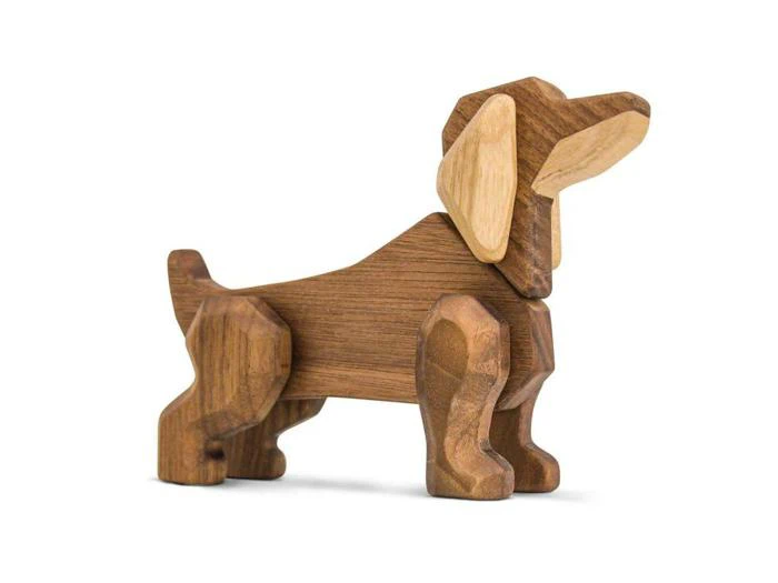 En hundefigur af træ med enkle, geometriske former og lyse og mørke trætoner, der står på alle fire ben mod en hvid baggrund.