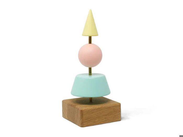 En geometrisk skulptur med en firkantet træbase, toppet af en lyseblå trapez, en pink kugle og en gul kegle, som alle er stablet lodret på en metalstang.