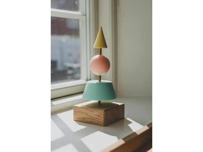 Et stykke trælegetøj med geometriske former - en gul kegle, en pink kugle og en blå cylinder - stablet på en stang, står i vindueskarmen i naturligt lys.