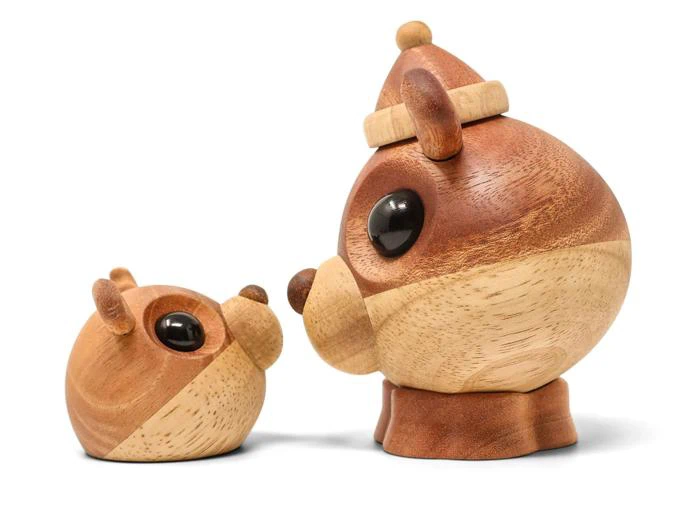 To bjørnefigurer af træ med store sorte øjne, den ene sidder oprejst og den anden ligger ned, begge lavet af lyst og mørkt træ med runde former og enkle træk.