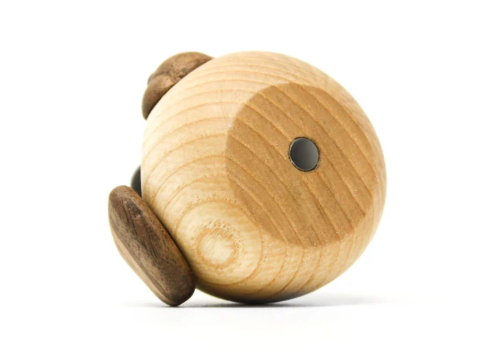 En lille, rund trægenstand med en lysere trækrop og mørkere trædetaljer, der muligvis ligner en minimalistisk dyrefigur, ligger på siden mod en hvid baggrund.