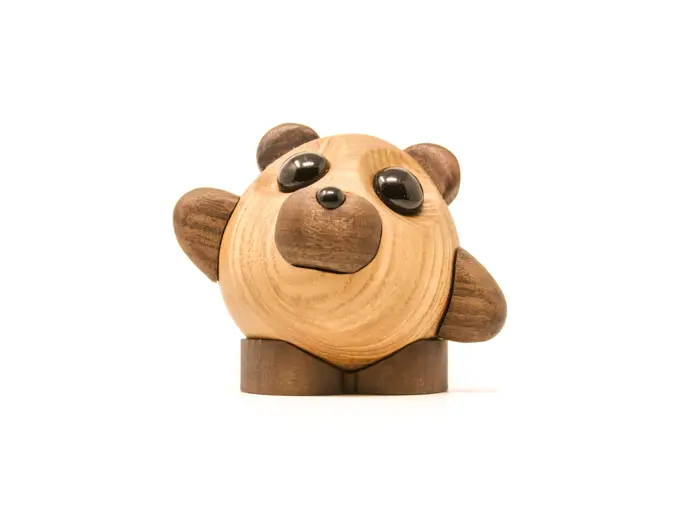 En lille, rund bjørnefigur af træ med store sorte øjne, to ører og løftet pote, som står på en hvid baggrund.