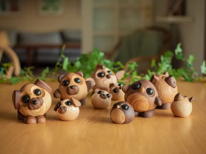 En gruppe dyrefigurer af træ med store sorte øjne, bl.a. hunde og bjørne, udstillet på et træbord med grønne planter i baggrunden.