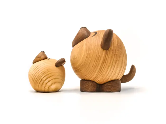 To runde dyrefigurer i træ med minimale detaljer, der ligner en lille og en stor hund, sidder på en hvid baggrund. Den store står, mens den lille hviler på siden.