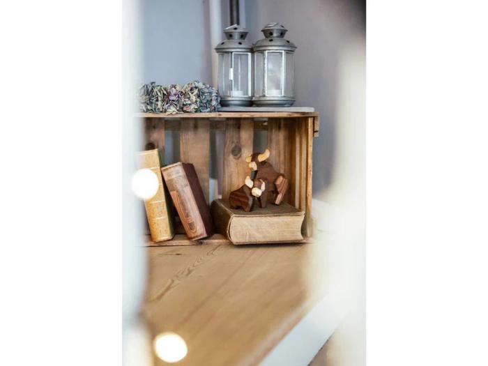 En trækasse rummer vintagebøger, en lille dyrefigur af træ, tørrede blomster og to sølvlygter, alt sammen anbragt på et trægulv.