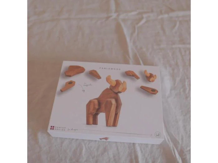En æske med dyrepuslespilsbrikker af træ, der er arrangeret på låget, og som viser, hvordan brikkerne samles til en stående dyrefigur, placeret på en lys stofoverflade.