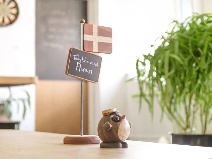 Et lille dansk flag på en stander med et skilt, hvor der står Tillykke med huen, ved siden af en fuglefigur i træ med studenterhue på, på et bord indendørs med en plante i baggrunden.