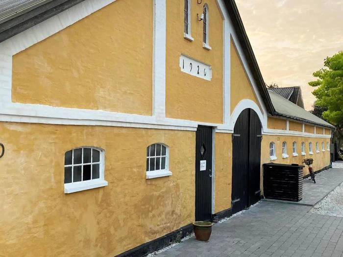 En lang, gul stukbygning med hvide kanter, sorte buede døre og små vinduer. Nummeret 1926 står over den store dør. En potteplante står nær indgangen.