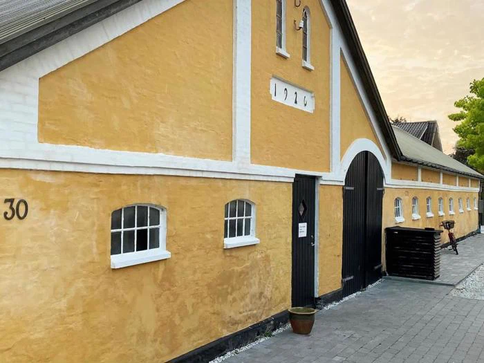 En lang gul bygning med hvid kant, sort buet dør og små vinduer. Nummeret 1924 står over døren, og nummer 30 står på venstre væg.