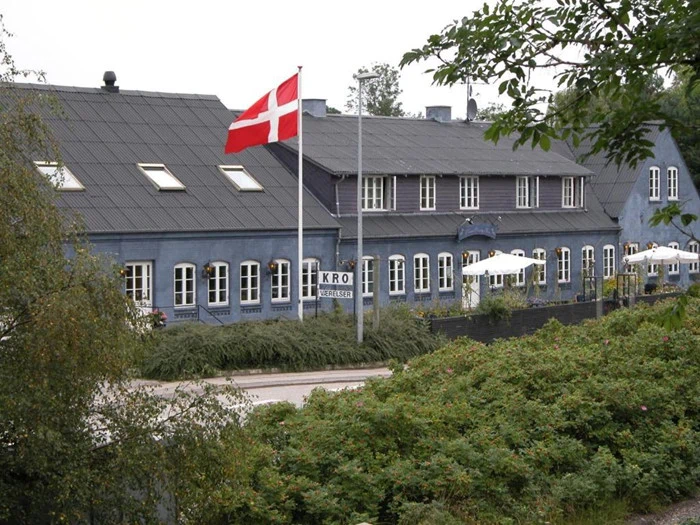 Et dansk flag vajer foran en blågrå bygning med buede vinduer og et KRO-skilt, omgivet af grønne områder og udendørs siddepladser med hvide parasoller.