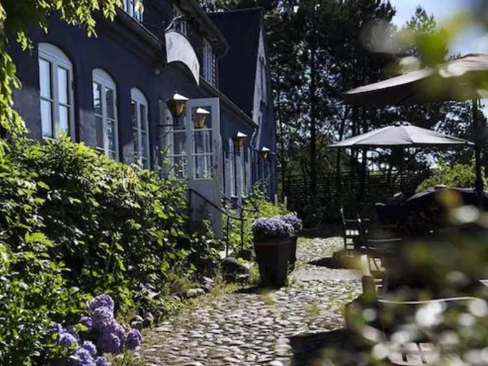 En charmerende brostensbelagt gårdhave ved siden af en blå bygning med buede vinduer, lilla blomster i potter og udendørs siddepladser i skyggen af parasoller.