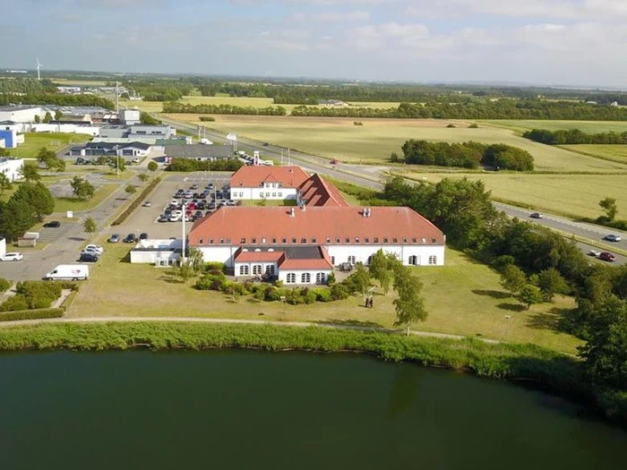 En stor bygning med rødt tegltag ligger ved en dam, omgivet af græs, træer, parkerede biler, veje og åbne marker under en delvis overskyet himmel.