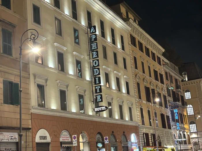 Natbillede af en bygade med oplyste bygninger med et stort lodret APHRODITE HOTEL-skilt på en af facaderne. Biler er parkeret langs fortovet.