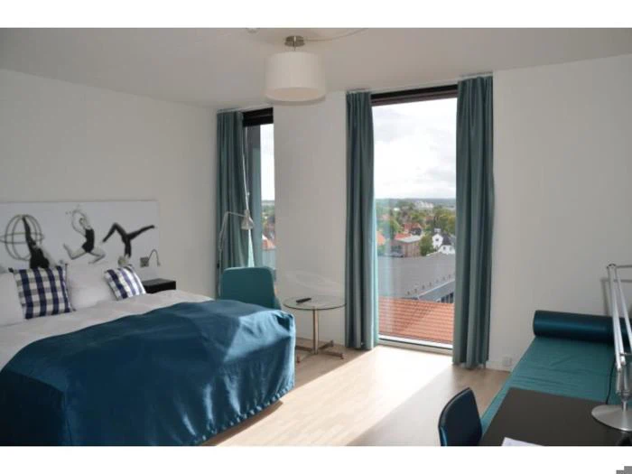 Et moderne hotelværelse med dobbeltseng, blåt sengetøj, ternede puder, stort vindue med blå gardiner, udsigt over byen, skrivebord og moderne indretning.