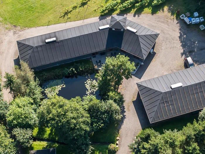 Luftfoto af moderne bygninger med sort tag, der omgiver en lille dam med grønne områder, træer og en grusbelagt indkørsel.