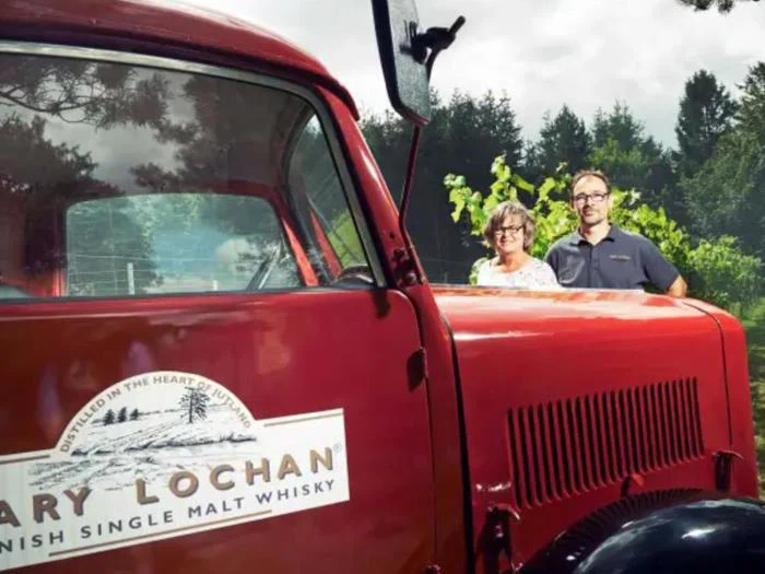 To personer står bag en stor rød lastbil med et "LIMAVADY LOCHAN IRISH SINGLE MALT WHISKY"-logo, omgivet af grønne områder og træer under en overskyet himmel.