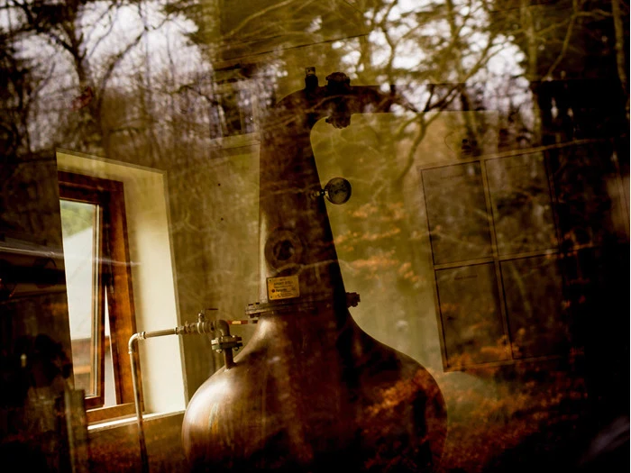 Et stort, gammelt kobberbrænderi ses gennem et vindue med refleksioner af træer og skov udenfor, hvilket blander indendørs og udendørs scener.