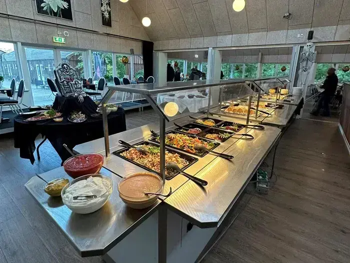 En buffetopstilling i et lyst spiseområde med bakker med forskellige salater, toppings og saucer med borde og stole i baggrunden.