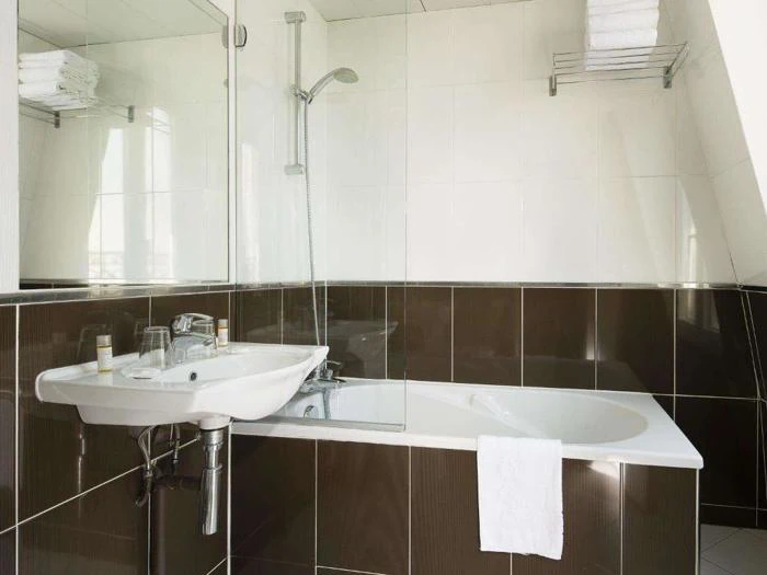 Et moderne badeværelse med badekar, bruseafskærmning i glas, væghængt håndvask, spejl og hylder med pænt foldede hvide håndklæder. Mørkebrune og hvide fliser pryder væggene.