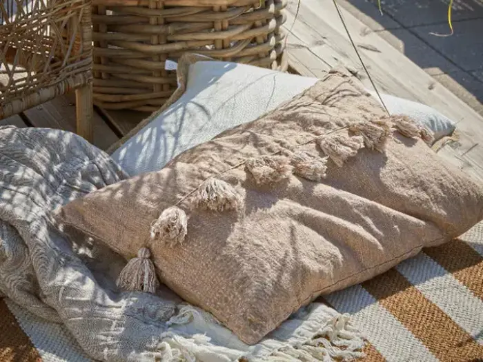 Beige tekstureret pude med kvaster på et tæppe ved siden af en flettet kurv på en udendørs træterrasse.