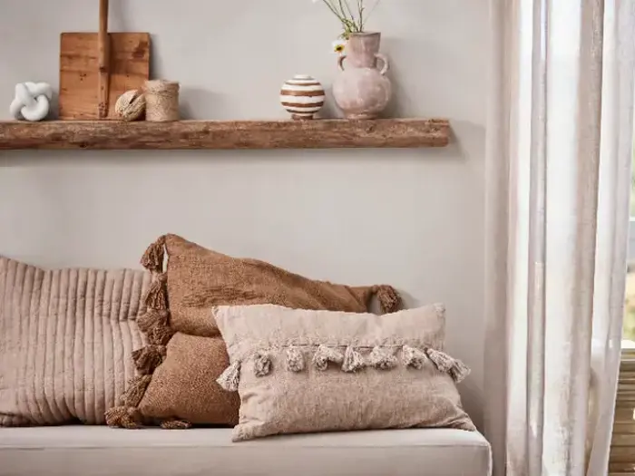 Neutralfarvet sofa med puder med tekstur og kvaster; rustik træhylde med vaser, en skål og dekorationsgenstande; sollyset trænger igennem de gennemsigtige gardiner.