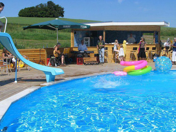 Et band spiller musik ved en pool med en blå rutsjebane, farverige poolflåd og folk, der slapper af udendørs på en solrig dag.