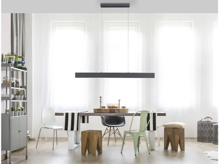 En moderne spisestue med et træbord, forskellige stole og skamler, en sort pendel og store lyse vinduer i baggrunden.