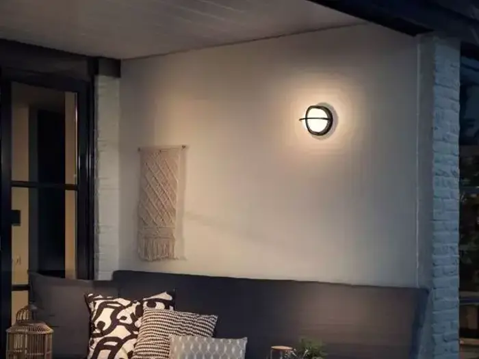 En hyggelig udendørs terrasse med en mørk, polstret bænk, mønstrede puder, en lille plante og en tændt væglampe ovenover, som oplyser den hvide murstensvæg og en lille macramé-dekoration.