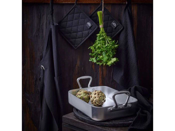 Et metalfad med to artiskokker står på en mørk bordplade. Ovenover hænger et bundt frisk persille ved siden af sorte grydelapper og et forklæde op ad en mørk trævæg.