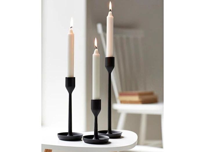 Tre tændte hvide stearinlys i moderne sorte lysestager i forskellige højder er anbragt på et hvidt bord. En hvid stol og stablede bøger er slørede i baggrunden.