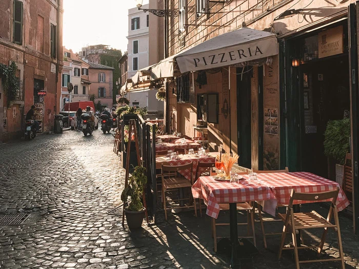 Udendørs borde med rødternede duge på et pizzeria på en brostensbelagt gade i en solbeskinnet europæisk by med bygninger og scootere i baggrunden.