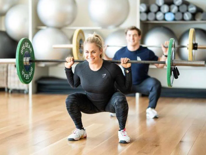 En kvinde og en mand laver squats med vægtstang i et fitnesscenter, begge smilende og iført sportstøj, med træningsbolde stablet i baggrunden.