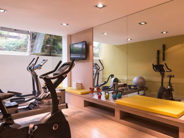 Et moderne motionsrum med motionscykler, kettlebells, et tv, en stabilitetsbold, store spejle og bænke med gule puder nær et vindue.