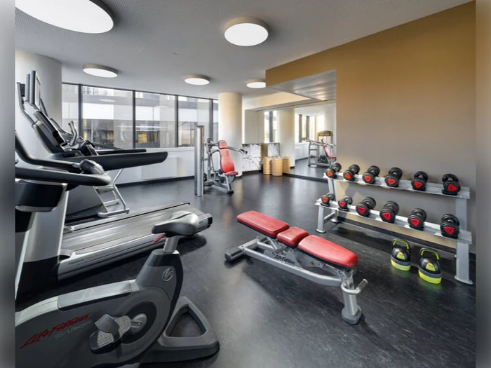 Moderne fitnesscenter med løbebånd, en stationær cykel, vægtstativer, håndvægte, røde bænke og store vinduer, der lukker naturligt lys ind.